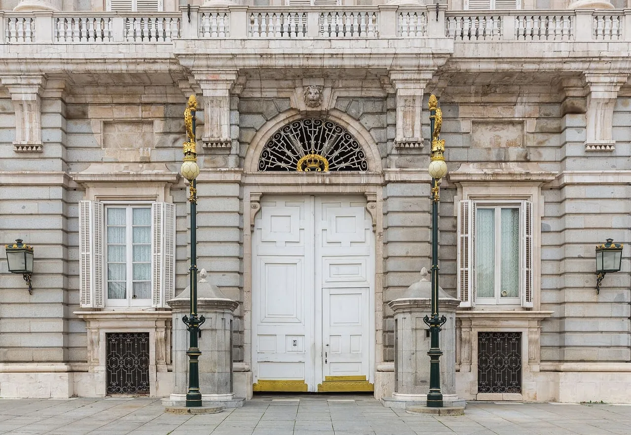 A white double door with a fan-shaped iron grille and gilded lamp posts in the grey stone wall of the Royal Palace of Madrid.