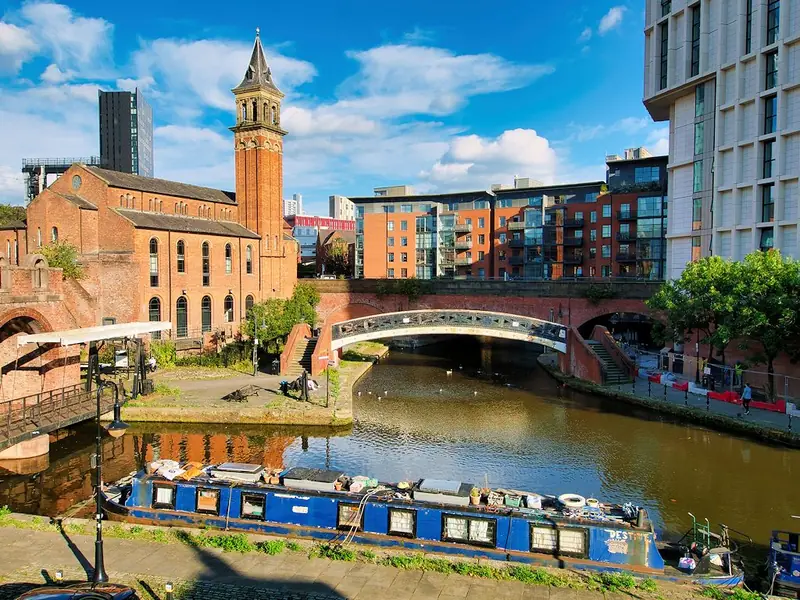 A canal basin with a moored narrowboat, a red-brick building with a clock tower, an arched bridge and modern apartment blocks behind.
