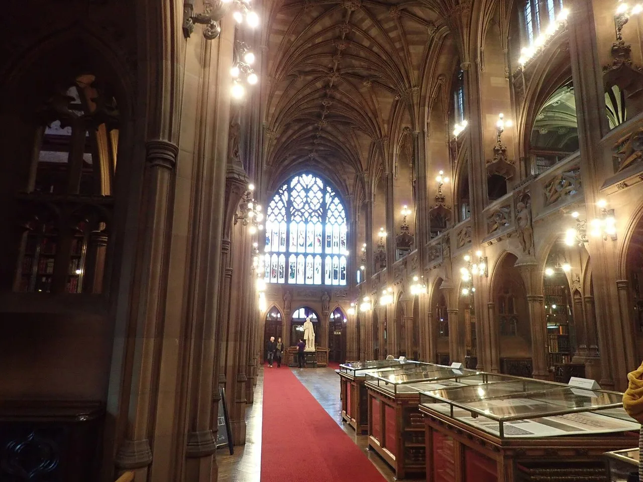 A long Gothic reading room with a vaulted ceiling, a pointed stained-glass window at the far end, a red carpet and glass display cases.