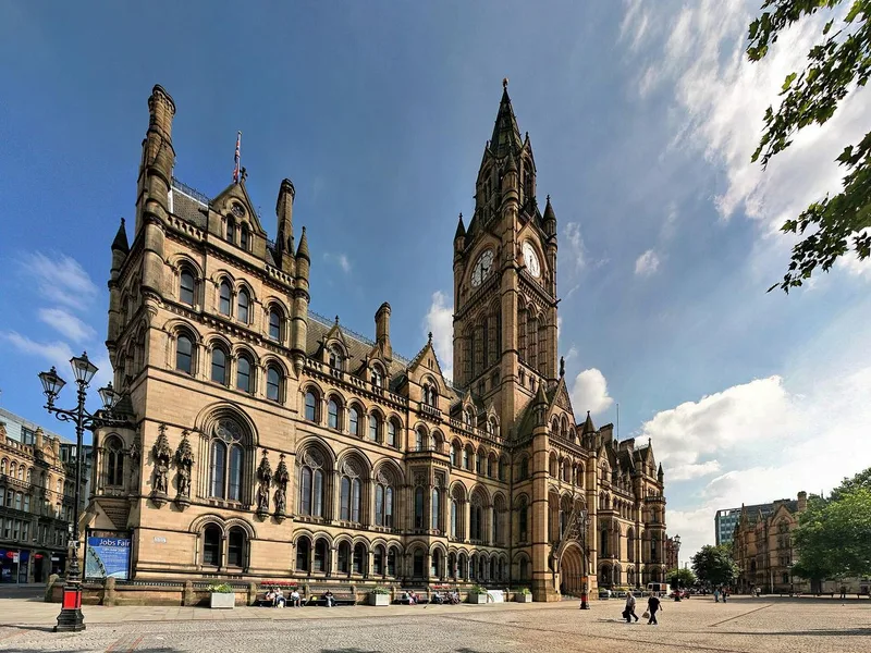 The Victorian Gothic Manchester Town Hall with its tall clock tower and pinnacled stone facade on a sunny day, seen from a paved square.
