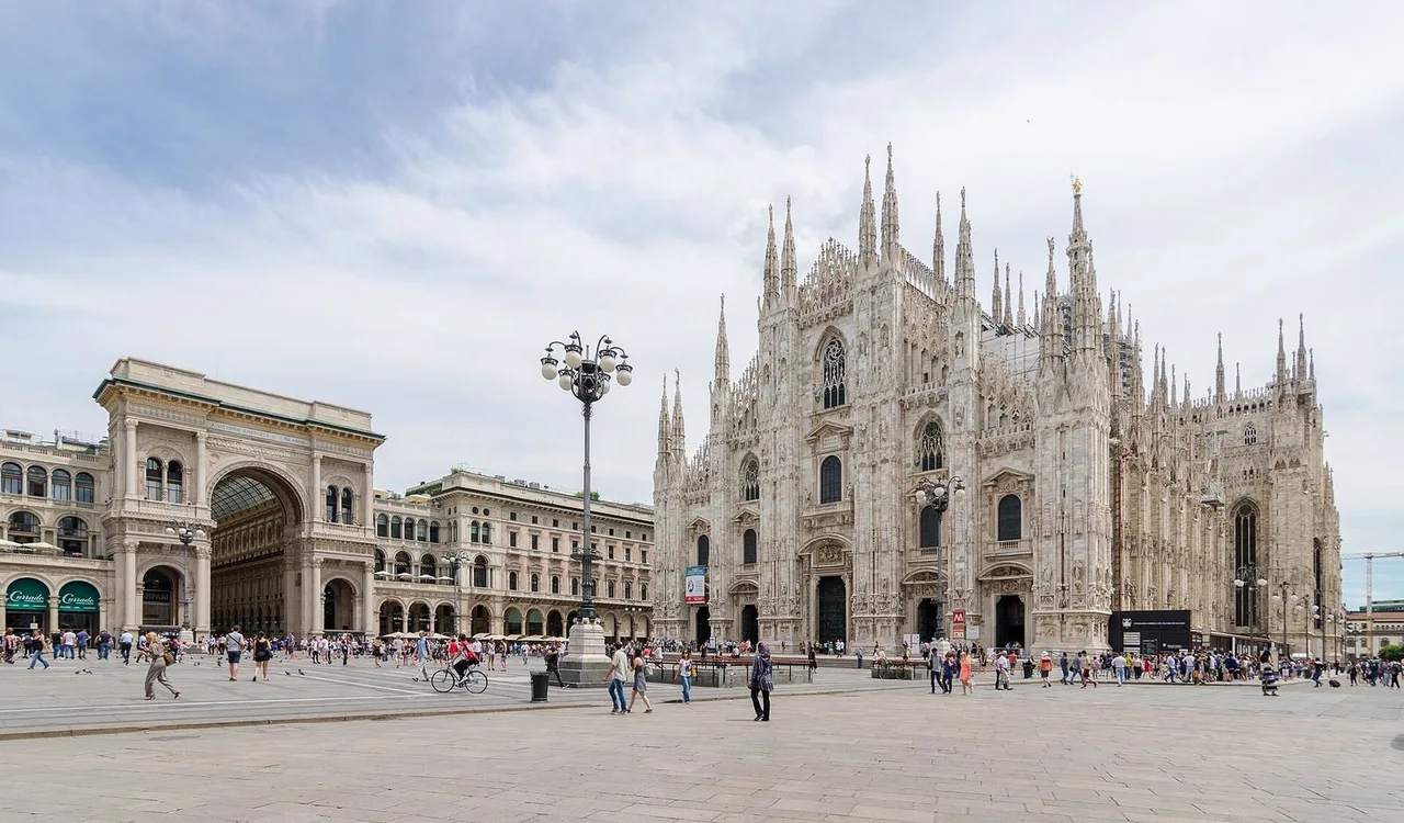 Milan Cathedral with its pale marble spires beside the arch of the Galleria Vittorio Emanuele II, seen across Piazza del Duomo under a cloudy sky.