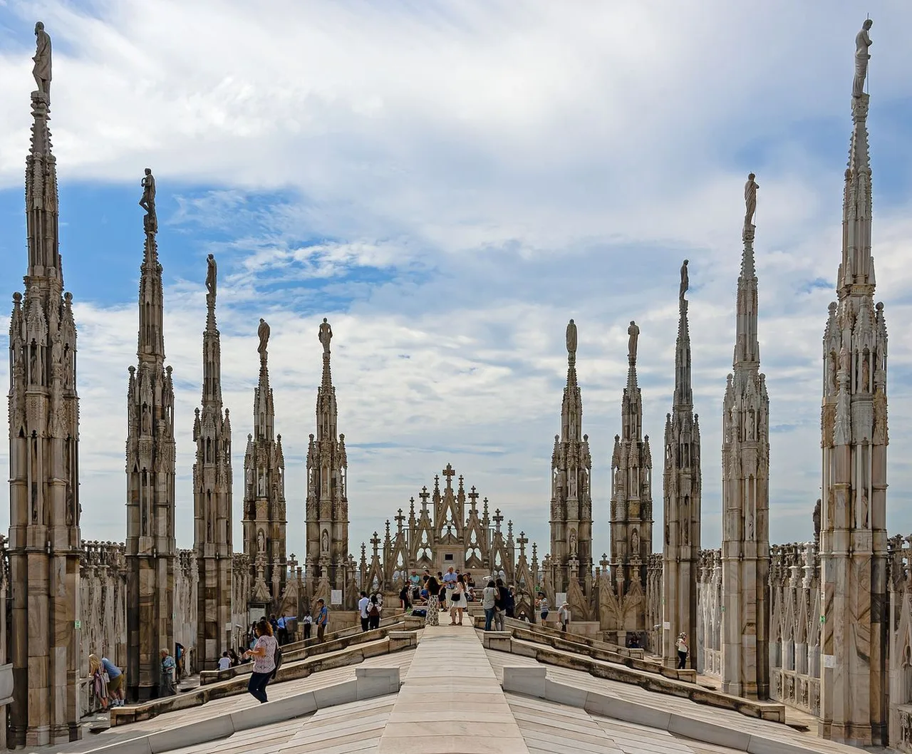 Rows of tall marble spires and statues along the walkable roof terraces of Milan Cathedral, with visitors far below under a partly cloudy sky.