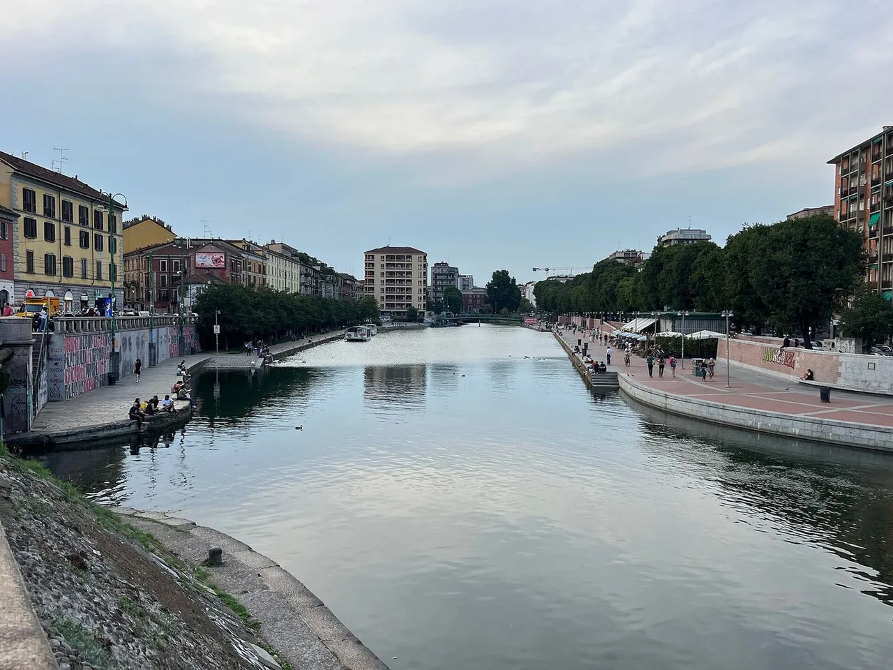 A wide canal between tree-lined stone quays and apartment buildings, with people sitting along the water on a cloudy evening.