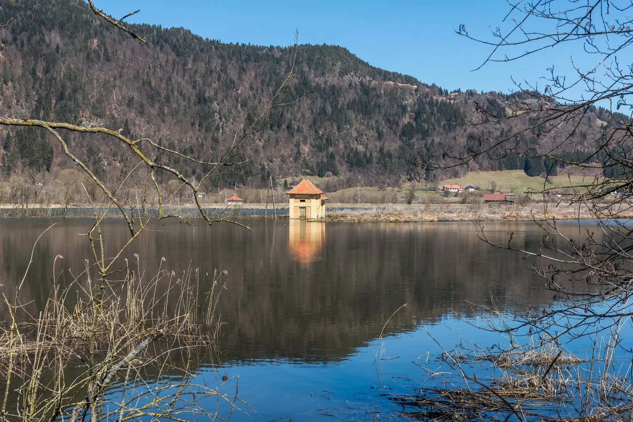 Small pump house with an orange roof standing in shallow water in the Bleistätter Moor, with wooded slopes behind