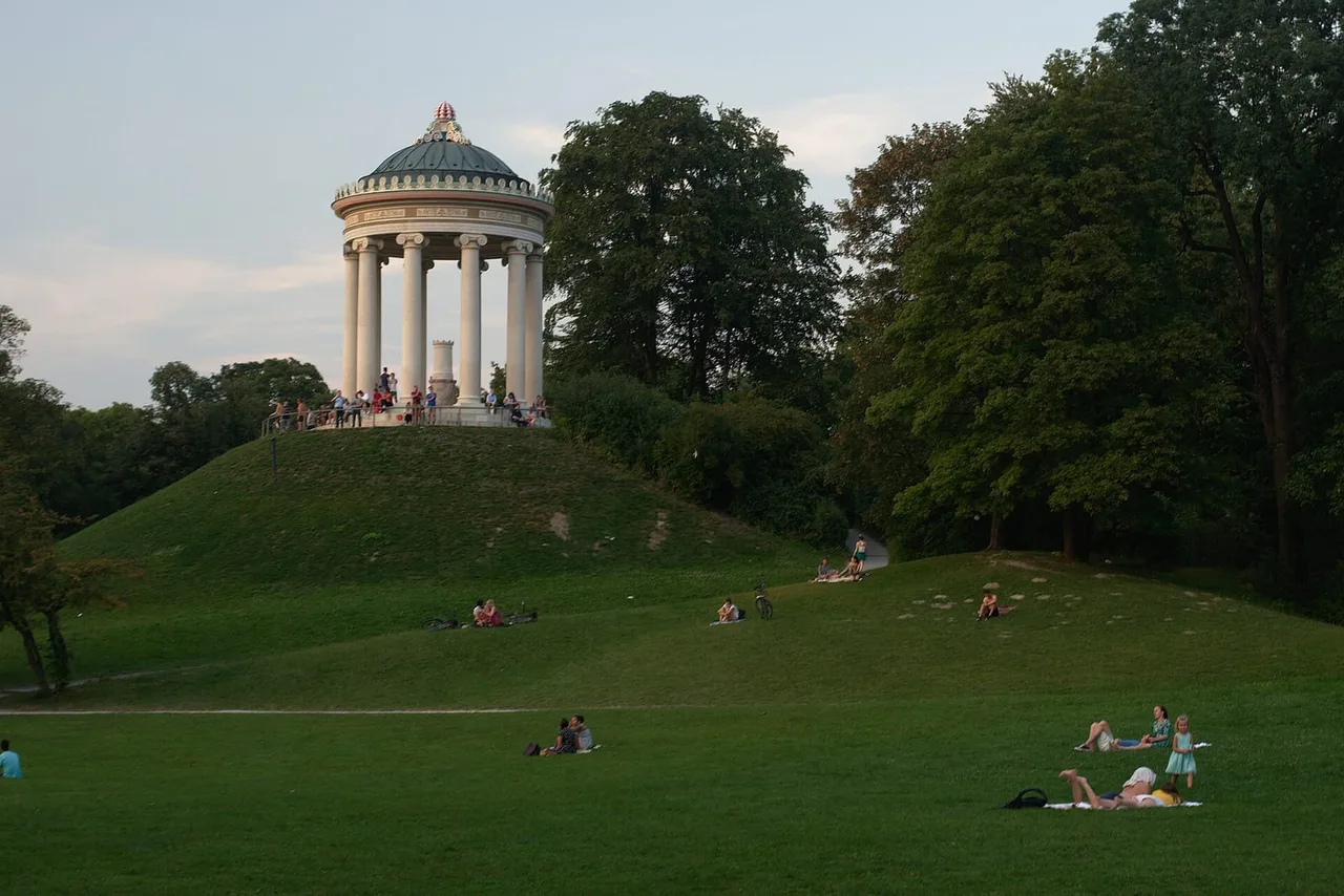 Round Greek-style temple with white columns on a grassy hill in a park, with people sitting on the slope and on the lawn below.