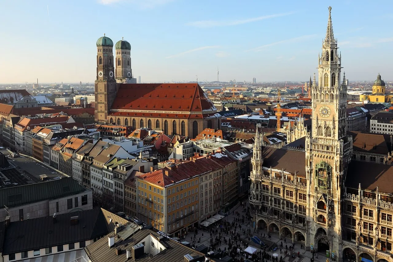 Rooftop view of Munich's old town with the twin green-domed towers of the Frauenkirche on the left and the tall tower of the Neues Rathaus on the right.