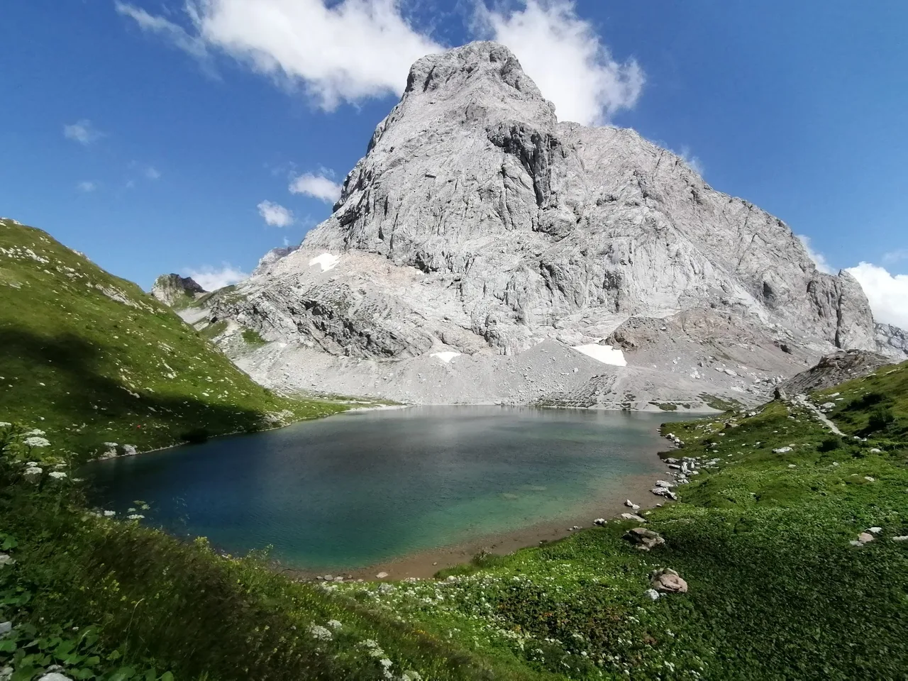 Turquoise lake at the foot of a large pale limestone peak, with green meadows and a few snow patches around the shore.
