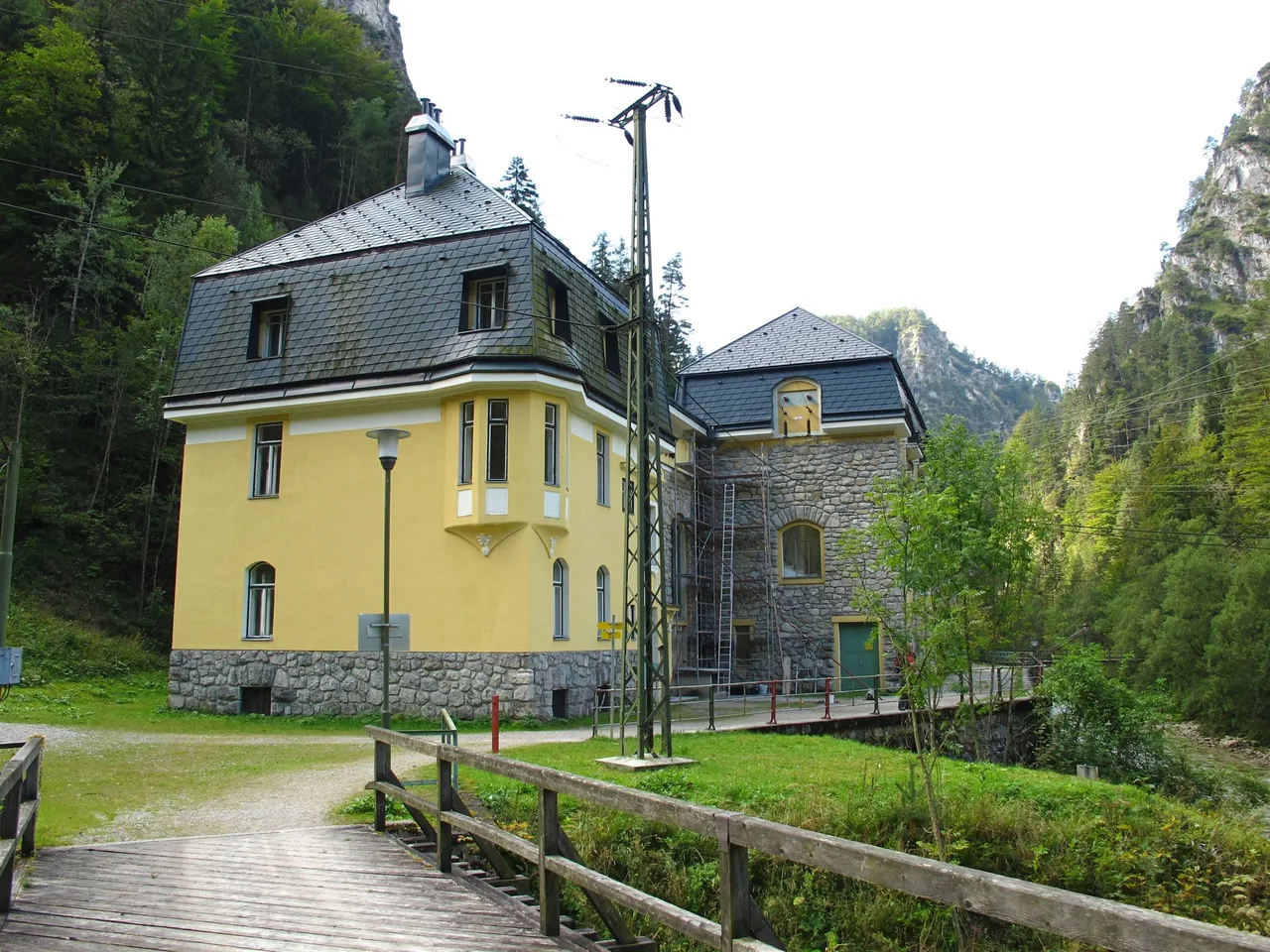 A yellow stone and slate-roofed power plant building stands beside a wooden footbridge and a fence, with forested rock walls behind it.