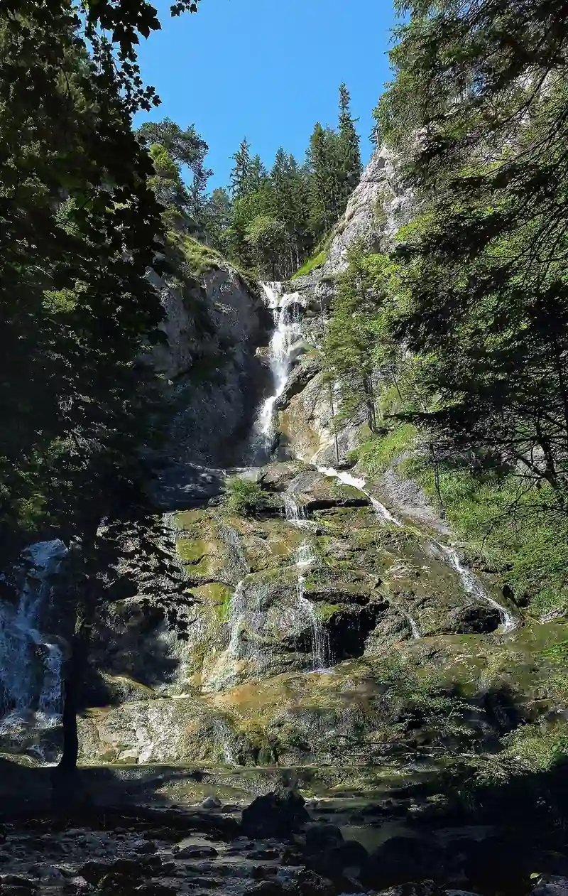 A waterfall runs in several steps down a mossy rock face in a shaded gorge under a blue sky.
