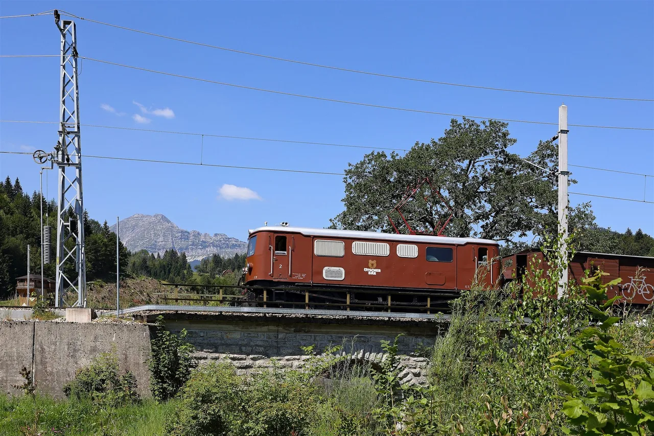 A red-brown electric locomotive with carriages crosses a bridge under overhead wires, with a mountain in the background.