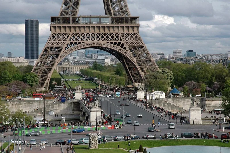 The base and first platform of the Eiffel Tower seen from Trocadéro, with the Pont d'Iéna, crowds, cars and the Champ de Mars lawns beneath its arch.