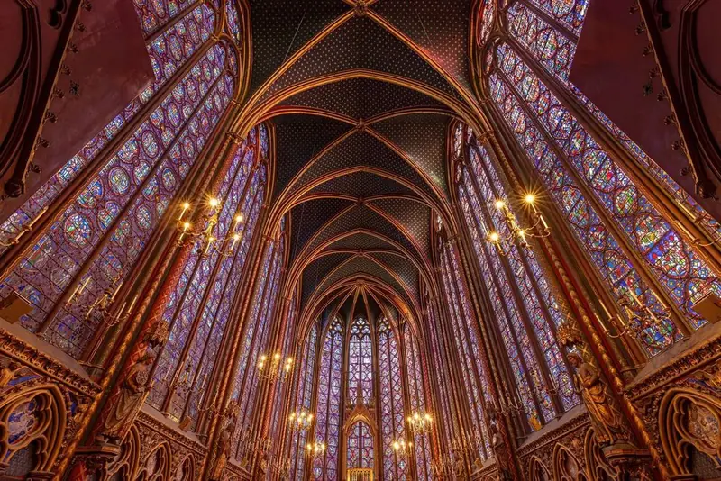 The upper chapel of the Sainte-Chapelle, with tall stained-glass windows on both sides, a star-studded vaulted ceiling and hanging candle chandeliers.