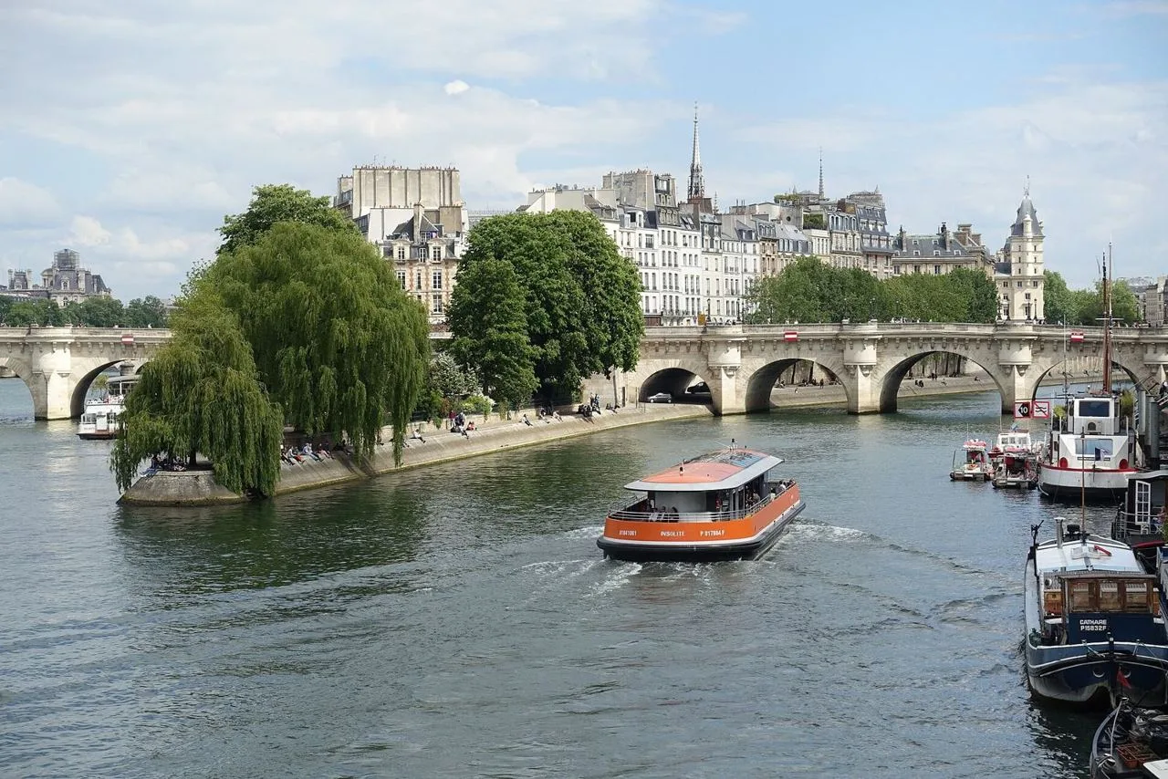 A tour boat on the Seine passing the willow-covered tip of the Île de la Cité, with the arches of the Pont Neuf and Paris apartment buildings behind.