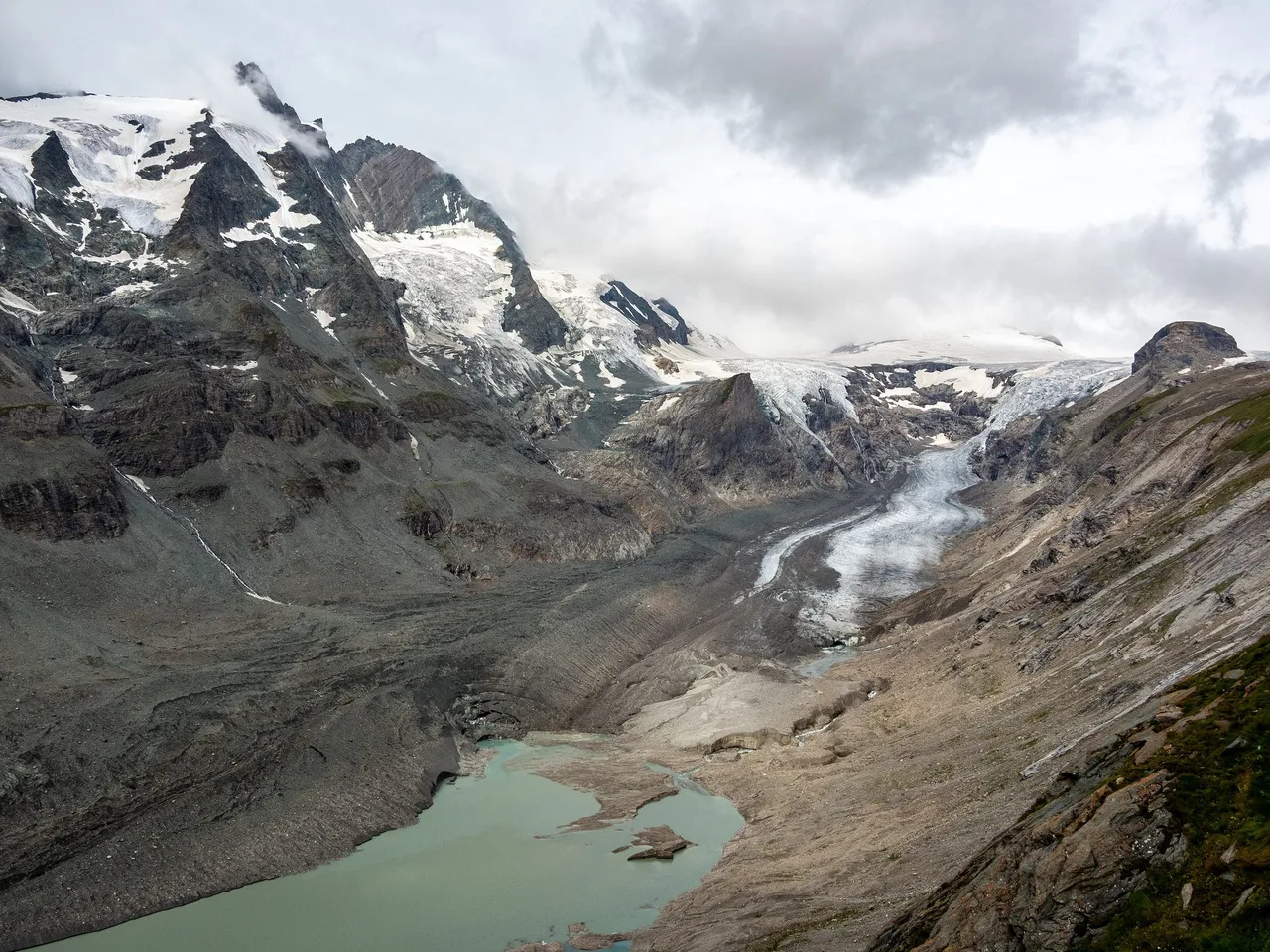 View down a glacier valley with a debris-covered ice tongue, a grey-green lake in the foreground and cloud-wrapped peaks of the Grossglockner group.