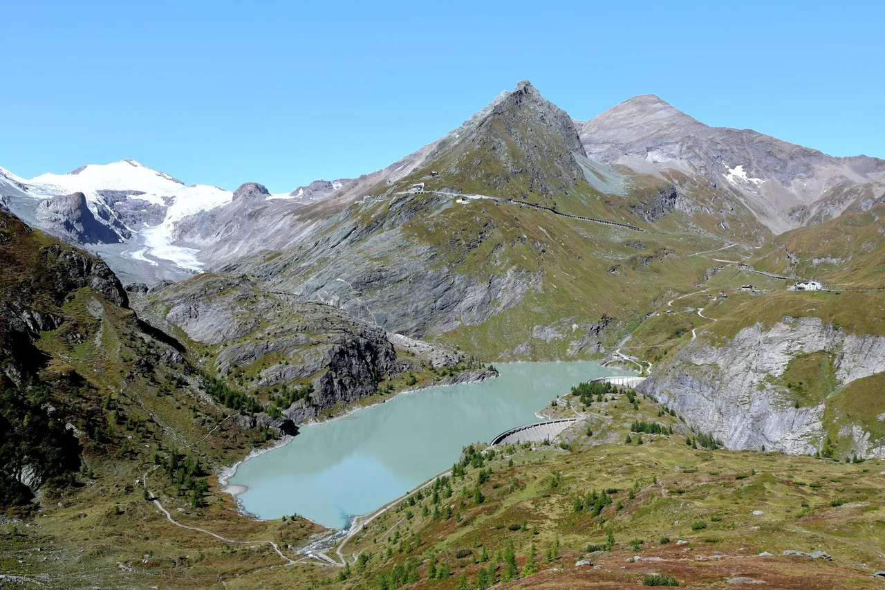Pale turquoise reservoir behind two concrete dams in a rocky high-mountain valley, with a small glacier on a ridge in the distance.