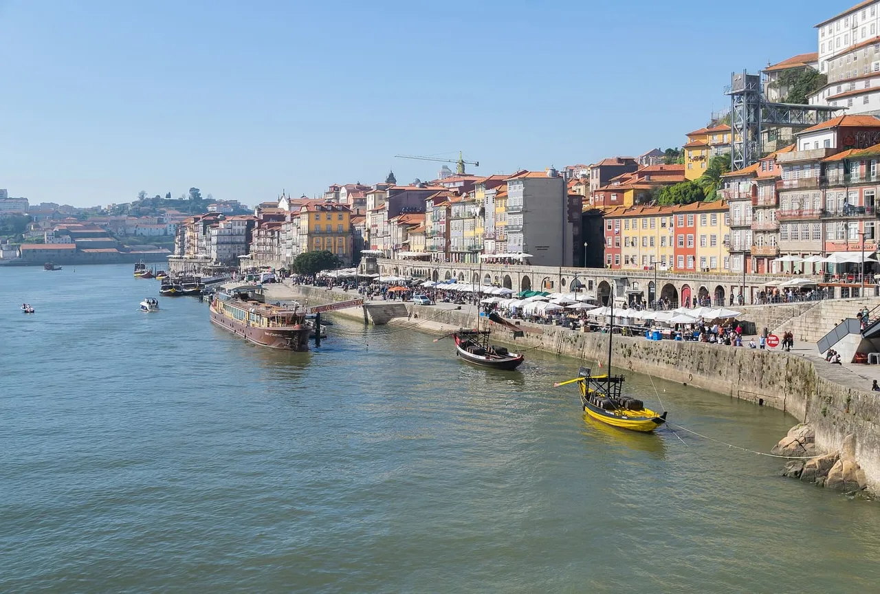 The Ribeira waterfront of Porto seen from above, with colourful houses on the hillside, café umbrellas along the quay and a yellow-hulled wooden boat moored on the Douro.