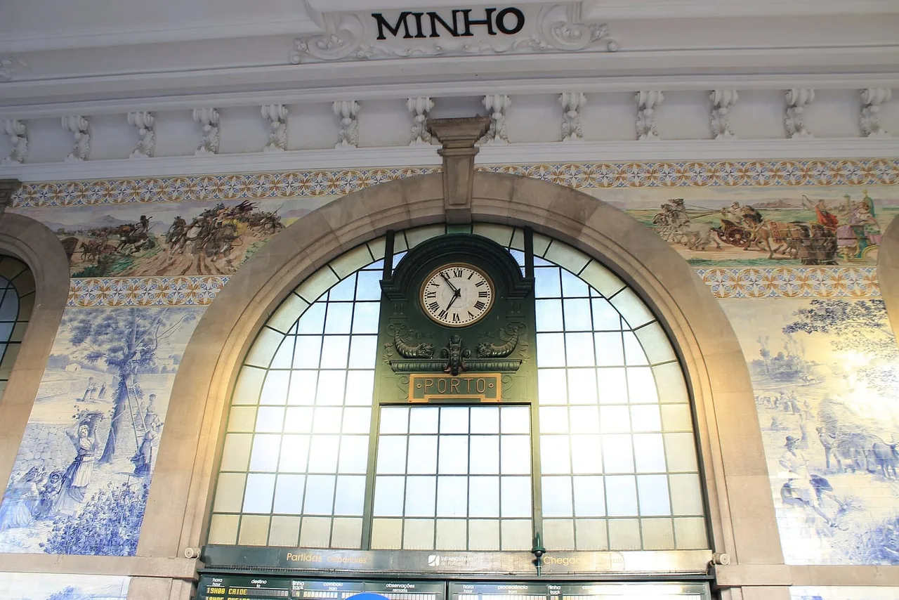 Blue and white azulejo tile panels with scenes of rural life and battles on either side of a large arched window with a clock in the São Bento station hall.