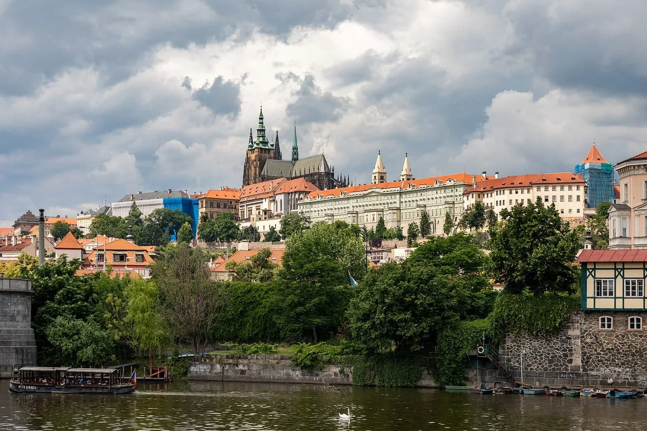 Castle hill in Prague seen across the river, with the spires of St Vitus Cathedral above long orange-roofed palace buildings under heavy clouds.
