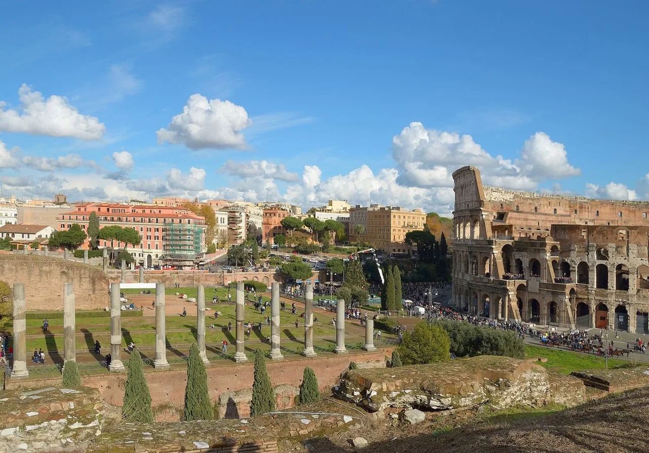 The Colosseum seen across the Roman Forum from the Via Sacra, with a row of ancient columns, pine trees and the city skyline behind.