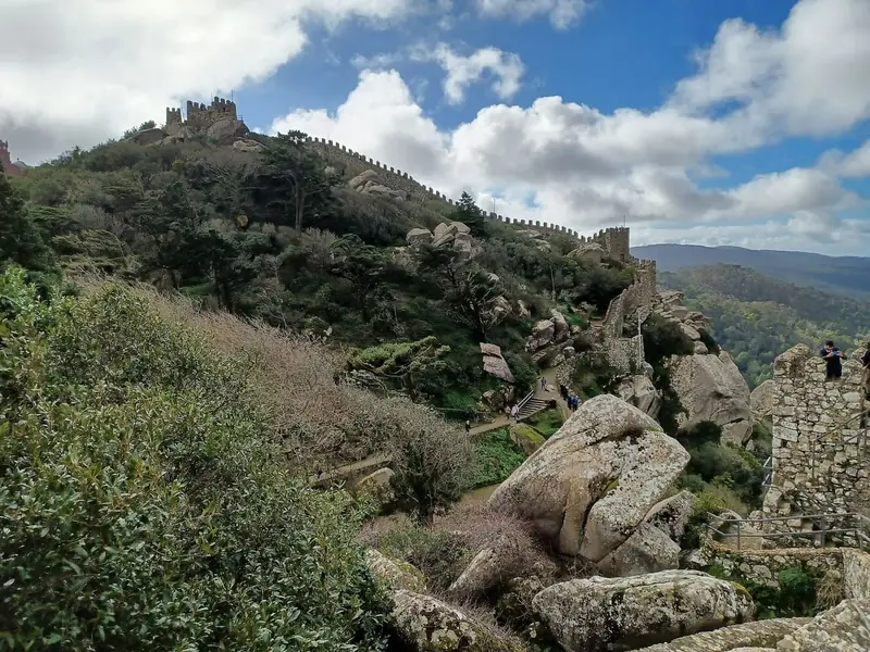 Battlemented walls of the Moorish Castle running along a rocky ridge covered in scrub under a partly cloudy sky, with a path and a few walkers.