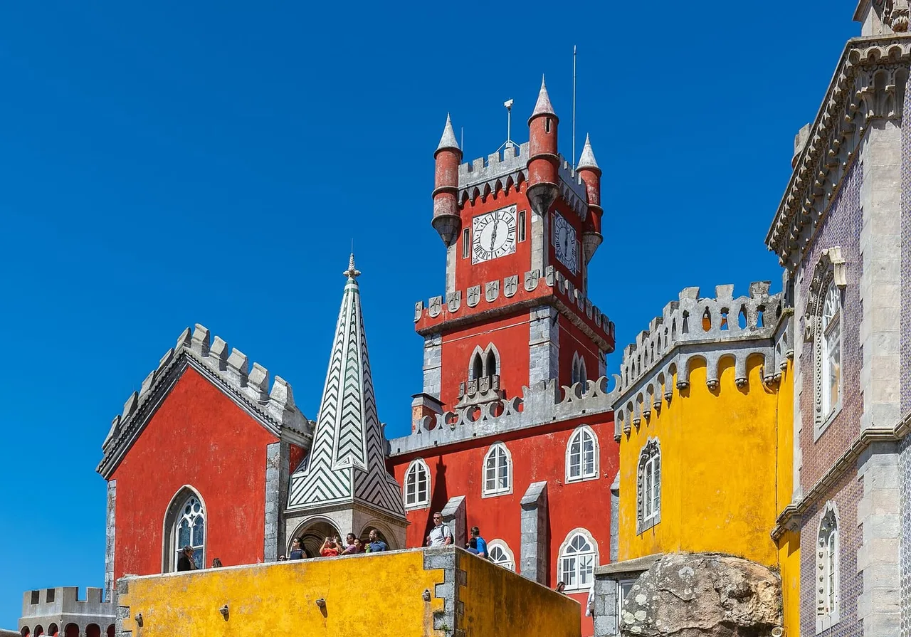 The red clock tower, yellow walls and striped green and white spire of Pena Palace against a clear blue sky, with a few visitors on a terrace.