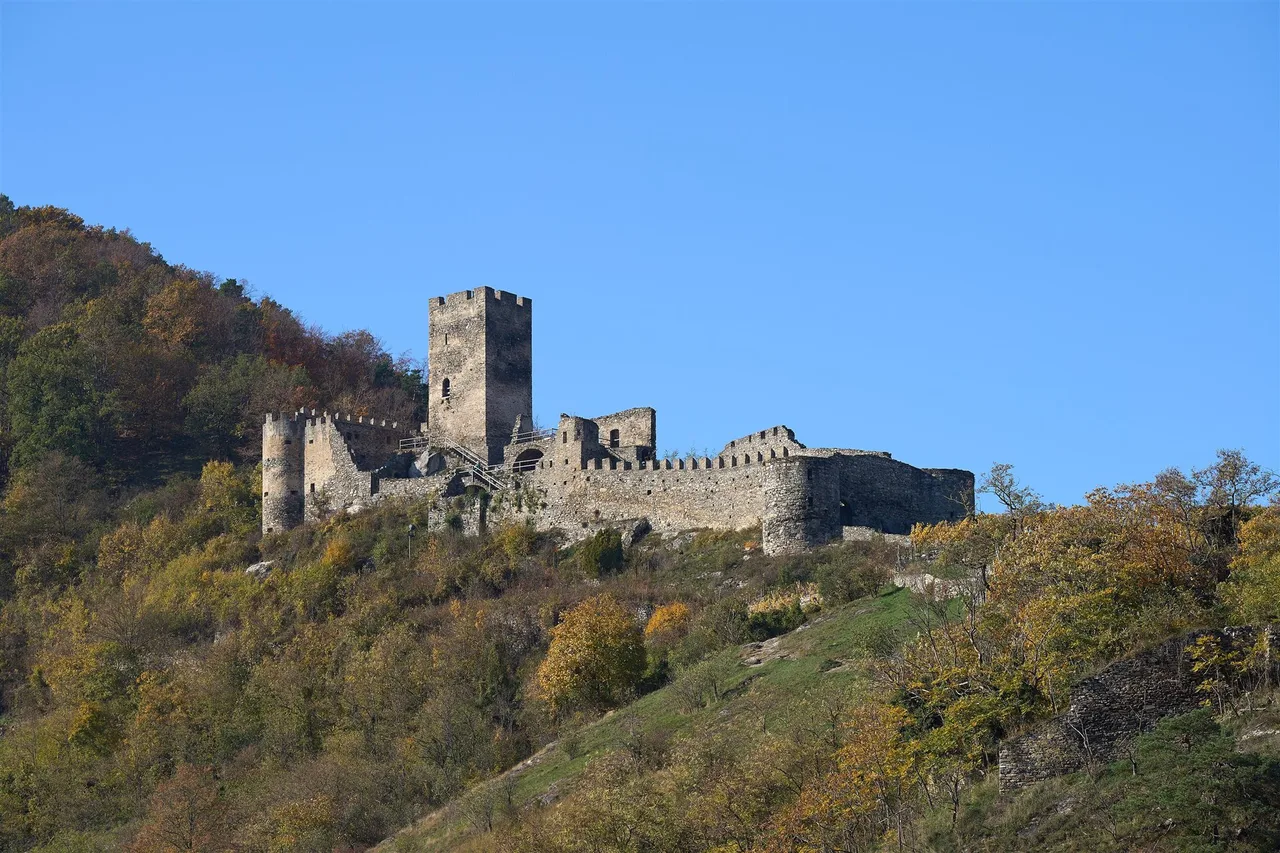 A castle ruin with a square tower and crenellated walls on a wooded slope in autumn colours under a clear sky.