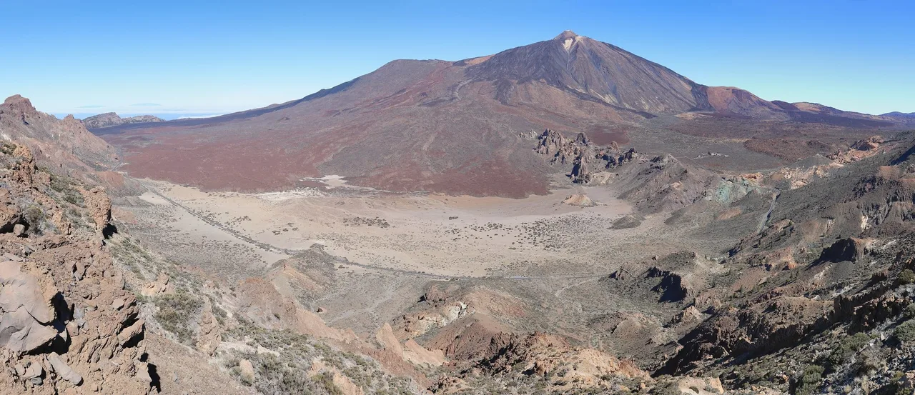 Wide view of the Teide cone rising above a pale caldera floor with reddish lava slopes and the Roques de García rock towers under a clear blue sky.