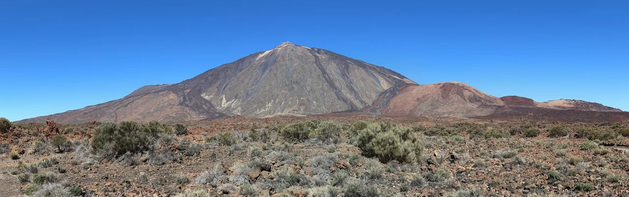 Panorama of the Teide cone beyond a rounded pale cone, Montaña Blanca, with scrub and lava boulders in the foreground.