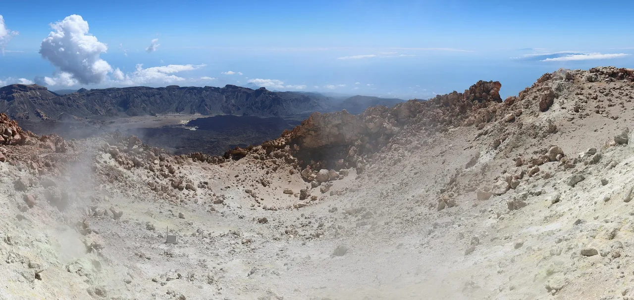 The pale, sulphur-stained rim of the Teide summit crater with wisps of steam, the caldera wall and a distant coast in the background.