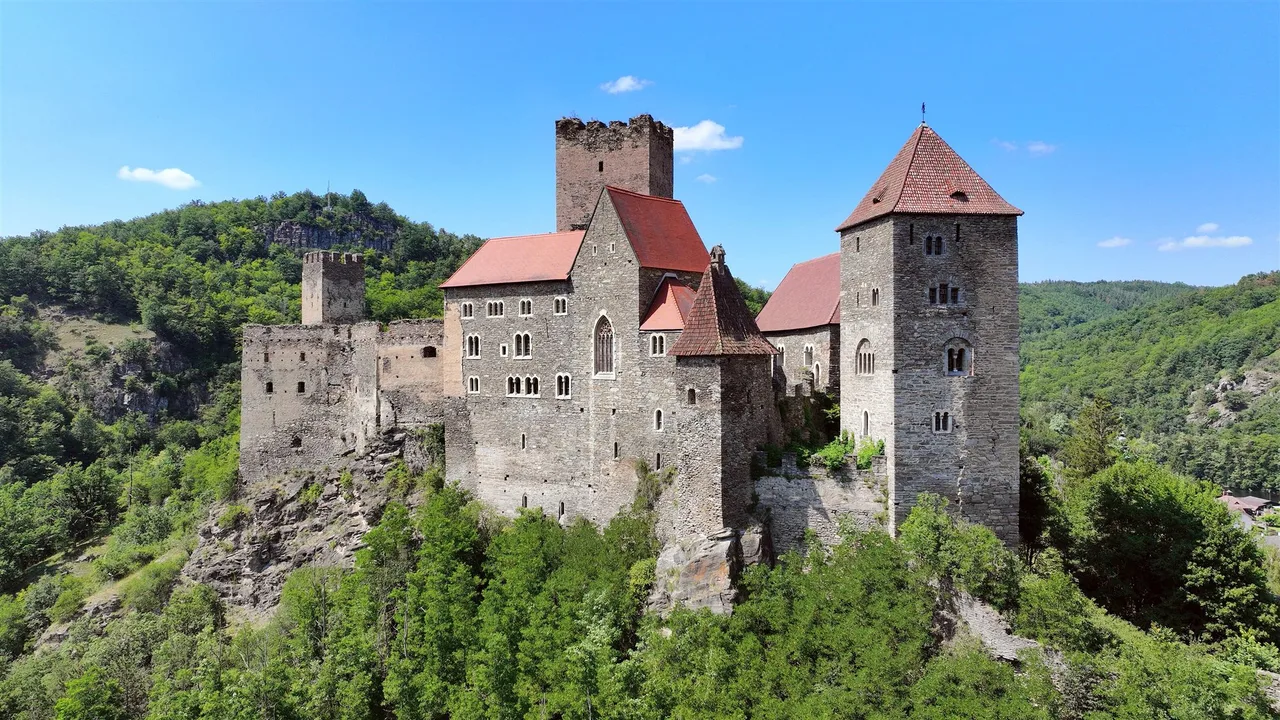 Hardegg Castle with its square towers and red roofs stands on a rock above wooded hills under a blue sky.