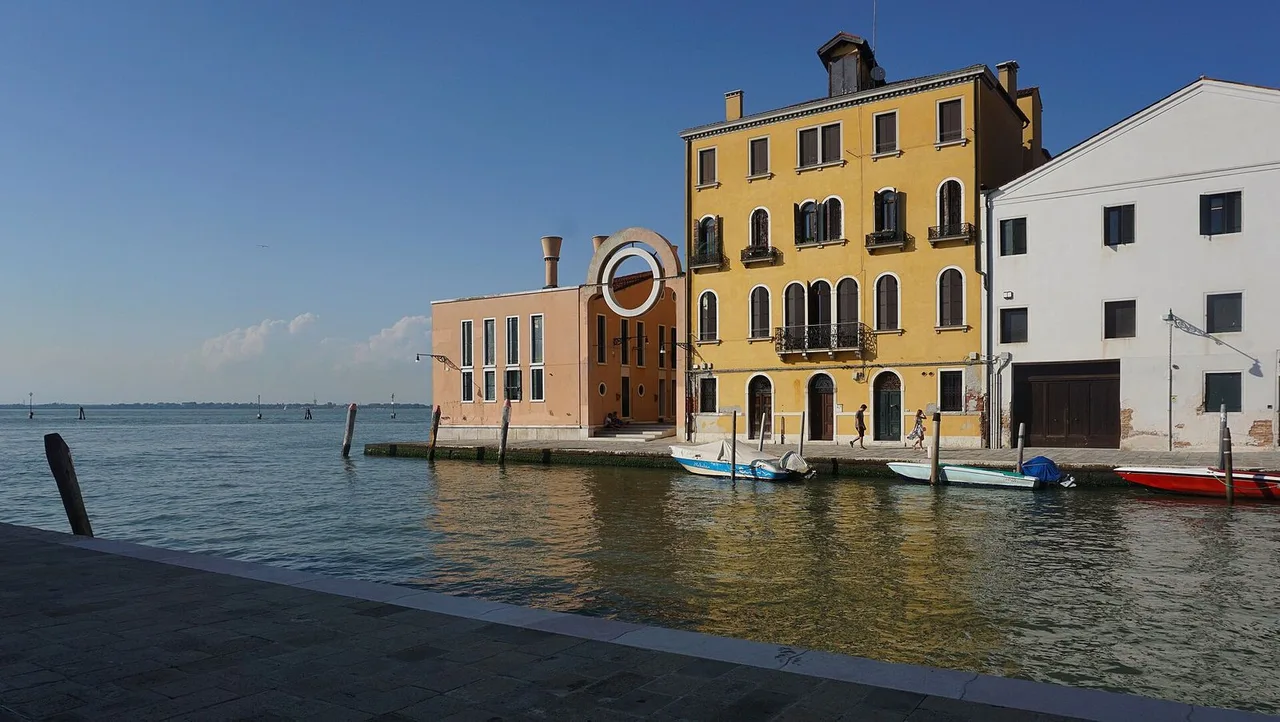 A yellow and a pink house with moored small boats beside a quay in northern Cannaregio, with open water and a blue sky behind.