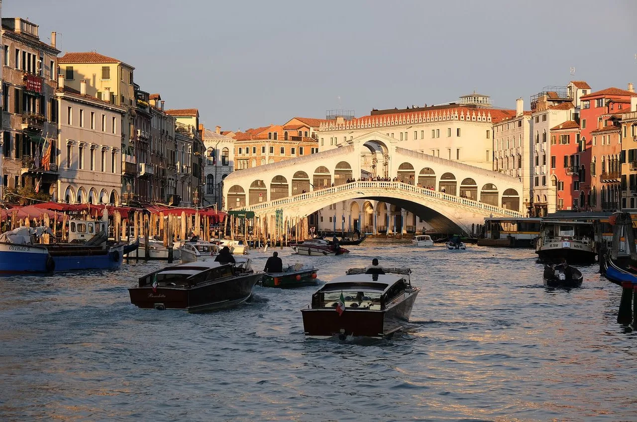 The white stone arch of the Rialto Bridge over the Grand Canal at warm evening light, with water taxis and boats in the foreground.