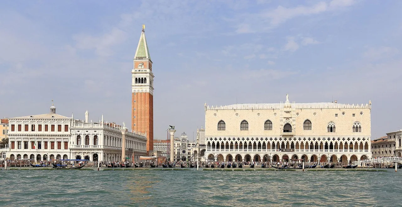 The red brick Campanile of St Mark and the pale arcaded Doge's Palace seen across the water of the lagoon, with the waterfront in front.