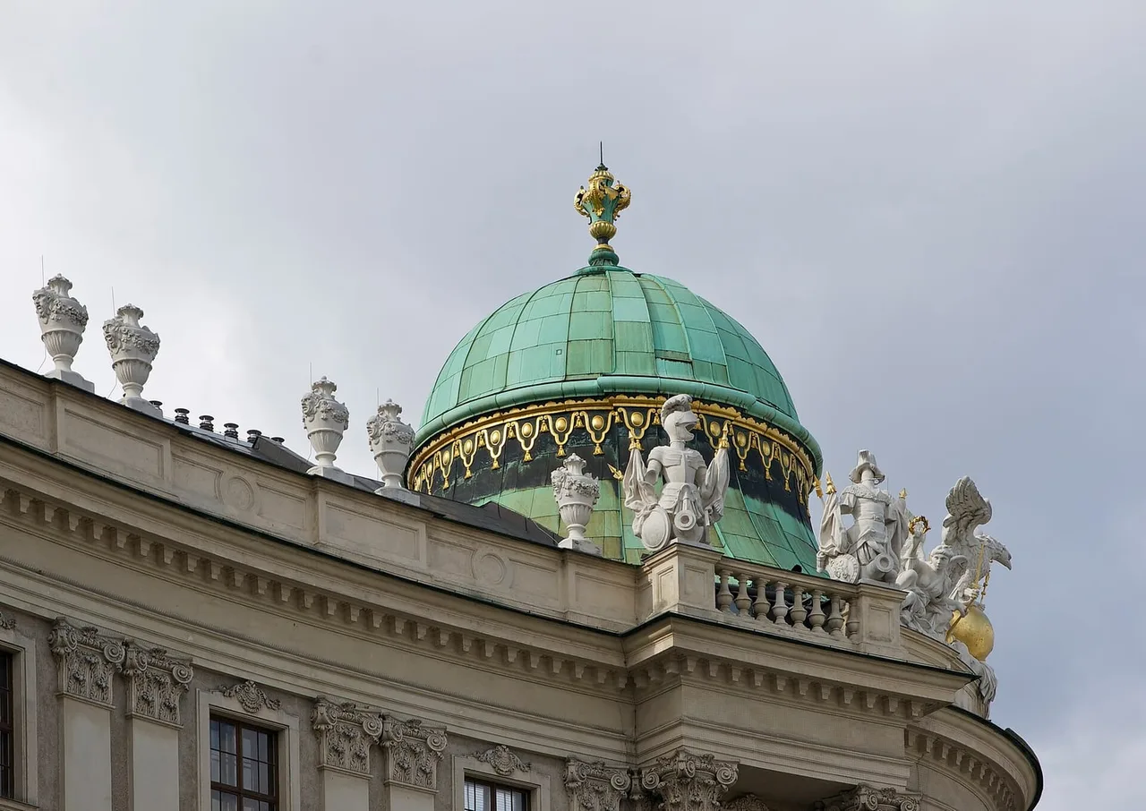 Green copper dome with gold trim and white stone statues and urns along the roofline of the Hofburg under a grey sky.