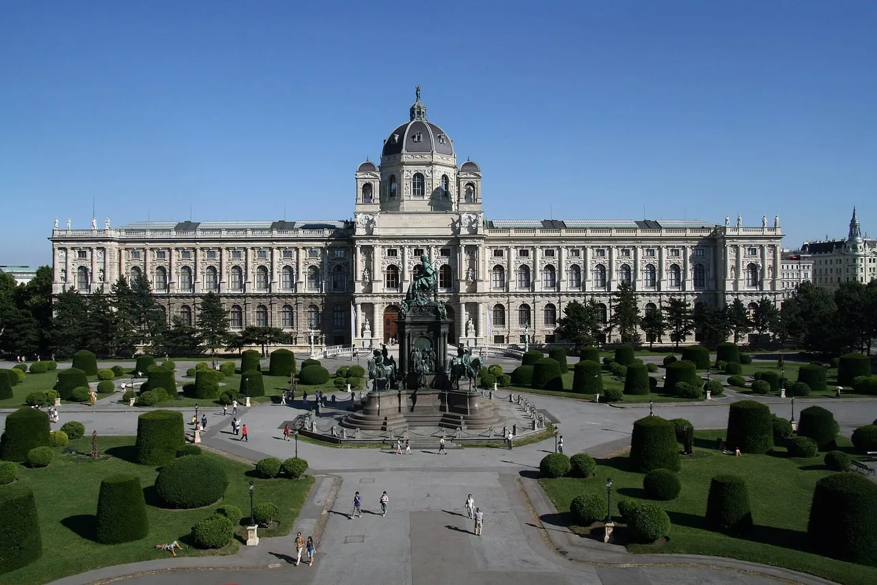 Large domed museum building on Maria-Theresien-Platz behind a monument and clipped hedges, with a few visitors on the paths.