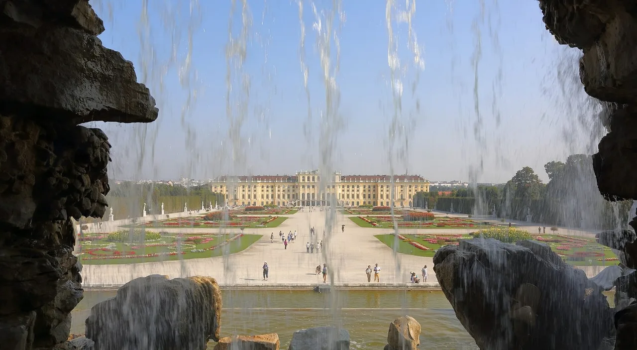 Yellow Schönbrunn Palace and its formal flower gardens seen through falling water at the Neptune Fountain, with visitors on the central path.