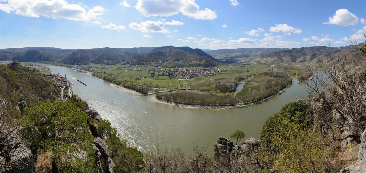 Panoramic view from Vogelberg over a wide bend of the Danube with villages, fields and wooded hills under a blue sky.