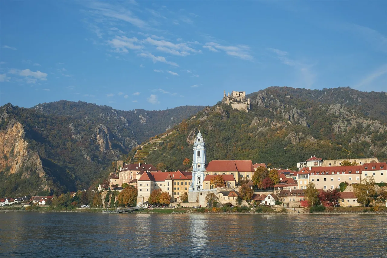 The town of Dürnstein on the Danube with a blue and white abbey tower, red roofs, a castle ruin on the ridge and trees in autumn colours.