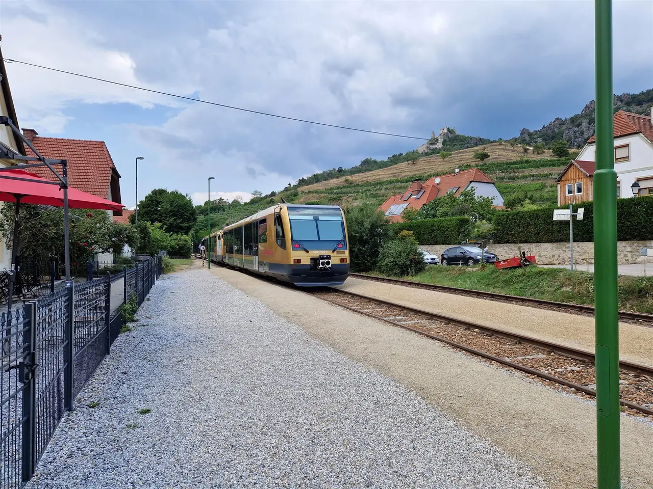A gold-coloured railcar approaches a small stop between houses, with vineyard terraces and a castle ruin on the hill behind.